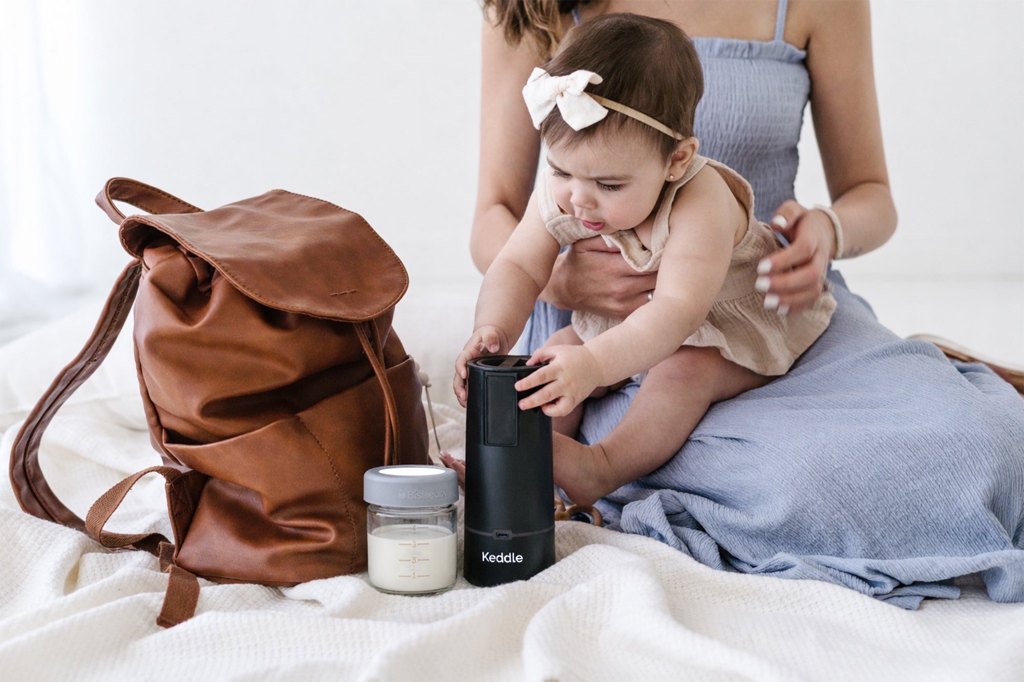 Mother and baby using the BisbeeBaby Keddle cordless portable bottle warmer alongside a glass breast milk storage jar, packed in a diaper bag for feeding on the go.