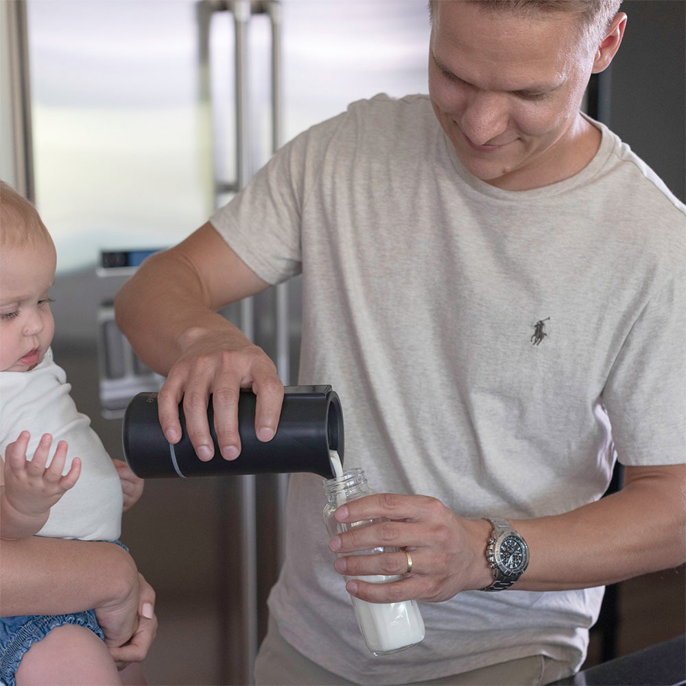 Man pouring milk from a travel bottle warmer into a glass with a baby sitting next to him.