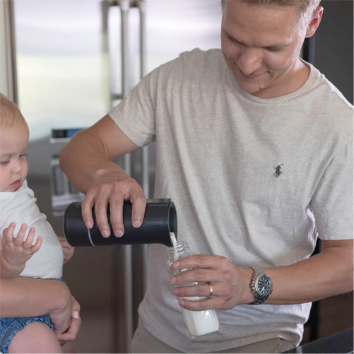 Man pouring milk from a travel bottle warmer into a glass with a baby sitting next to him.